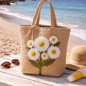 May include: A beige crochet tote bag with a daisy floral design. The bag has two handles and a bow detail. Sunglasses, a straw hat, and sunscreen are on a wooden bench near the beach.