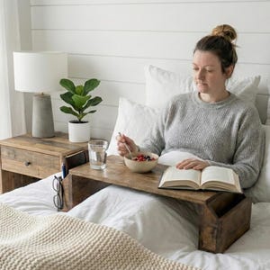 May include: A wooden bed tray with a book, bowl of fruit, and a glass of water. A woman is sitting on a bed with white linens, wearing a gray sweater. A side table with a lamp and a plant is visible.