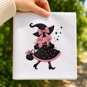 May include: A white fabric square featuring an embroidered design of a figure wearing a black witch hat with a pink bow, a black dress with pink polka dots and trim, pink hair, and black boots, carrying a black bag with pink accents and star motifs.