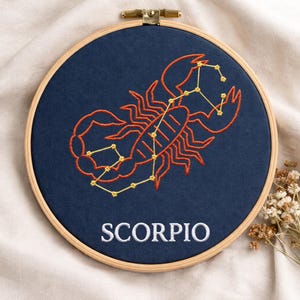 Peut inclure: Broderie du signe astrologique du Scorpion sur tissu bleu marine, dans un cercle en bois. Le motif présente un scorpion orange et une constellation avec des coutures jaunes, et le mot "SCORPIO" en blanc.