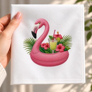May include: A white cloth napkin featuring an embroidered design of a pink flamingo pool float. The flamingo is surrounded by bright pink hibiscus flowers, green palm leaves, and a glass of yellow cocktail with a straw and lime slice.