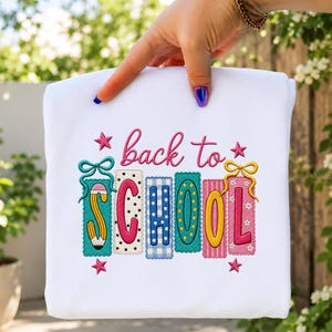 May include: A white t-shirt featuring an embroidered design with the text back to SCHOOL in pink cursive and block letters. The letters are decorated with colorful patterns, including a yellow pencil, polka dots, and gingham, accented by bows and stars.