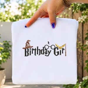 Puede incluir: Una camiseta blanca con texto bordado en negro que dice Birthday Girl. El diseño incluye un sombrero seleccionador marrón, gafas redondas negras con un rayo, una snitch dorada con alas y una varita marrón como elementos decorativos.