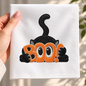 May include: A white square fabric napkin featuring an embroidered black cat with its tail raised, holding a large orange sign that reads BOO! in bold letters. The cat has wide white eyes and is centered on the fabric, showcasing detailed stitching.