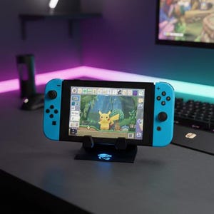 May include: A blue Nintendo Switch console displaying a video game with a Pikachu character. The console is mounted on a black stand with a glowing blue design. The background includes a keyboard and a monitor.