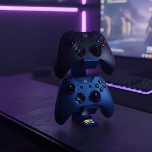 May include: A black and a blue Xbox controller are stacked on a blue stand. The controllers have buttons and joysticks. The stand has a gold emblem. The background is a dark room with a computer.