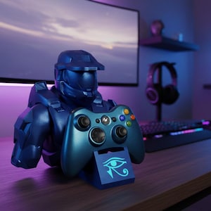 Può includere: Un controller di gioco blu è appoggiato su un supporto abbinato a forma di casco spartano blu. Il supporto presenta un simbolo stilizzato dell'occhio. Lo sfondo include un monitor del computer, una tastiera e delle cuffie.