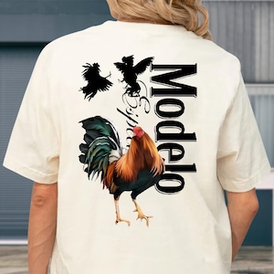 May include: Cream-coloured t-shirt with a colourful rooster graphic and black bird silhouettes. The word "Modelo" is printed vertically in black, with "Especial" in cursive. The shirt is a casual wear item, suitable for everyday use.