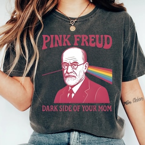 Peut inclure: T-shirt gris foncé avec l'inscription "PINK FREUD" en lettres roses. Le t-shirt présente un graphisme de Sigmund Freud avec un rayon arc-en-ciel émanant de son œil, et le texte "DARK SIDE OF YOUR MOM" en dessous.
