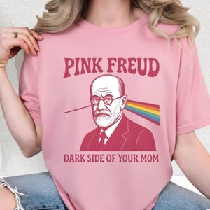 May include: A light pink t-shirt with the text "PINK FREUD" above an illustration of Sigmund Freud wearing glasses. A rainbow beam emanates from his head. The text "DARK SIDE OF YOUR MOM" is below the illustration.