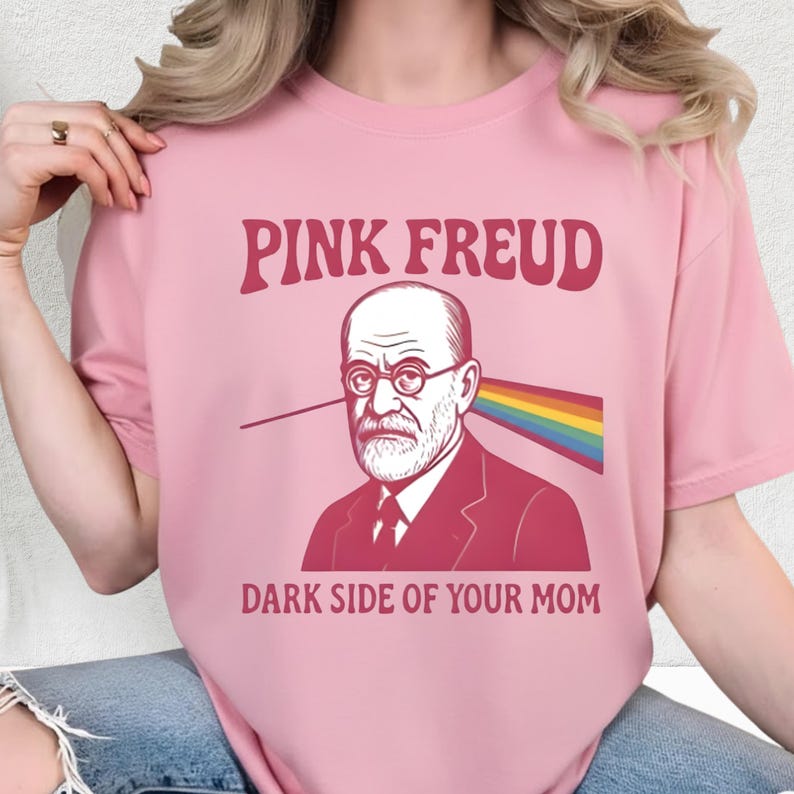 May include: A light pink t-shirt with the text "PINK FREUD" above an illustration of Sigmund Freud wearing glasses. A rainbow beam emanates from his head. The text "DARK SIDE OF YOUR MOM" is below the illustration.