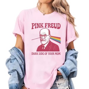 May include: A light pink t-shirt featuring a graphic of Sigmund Freud with the text "PINK FREUD" above and "DARK SIDE OF YOUR MOM" below. The graphic includes a rainbow prism effect.