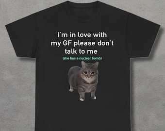 May include: Black t-shirt featuring a gray cat graphic. The text above the cat reads, "I'm in love with my GF please don't talk to me (she has a nuclear bomb)." The cat has gray and white fur.