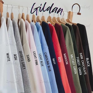 May include: A display of Gildan brand sweatshirts in various colors, including white, ash, sports grey, sand, light pink, light blue, Carolina blue, navy, red, maroon, military green, forest green, dark chocolate, and black. Each sweatshirt is labeled with its color.