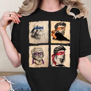 May include: Black t-shirt featuring four Renaissance-style portraits, each with a colored eye mask. The portraits are arranged in a 2x2 grid. The masks are blue, orange, purple, and red. The portraits are on a beige background.
