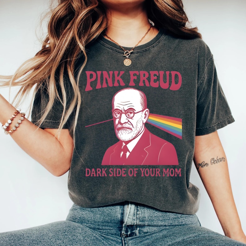 May include: Dark grey t-shirt with the text "PINK FREUD" in pink lettering. The shirt features a graphic of Sigmund Freud with a rainbow beam emanating from his eye, and the text "DARK SIDE OF YOUR MOM" below.