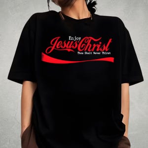 Könnte beinhalten: Schwarzes T-Shirt mit dem Schriftzug "Enjoy Jesus Christ" in roter, stilisierter Schrift, die an ein bekanntes Logo erinnert. Der Text "Thou Shalt Never Thirst" steht darunter.