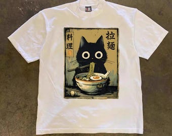 May include: White t-shirt featuring a graphic of a black cat eating ramen noodles from a bowl. The cat has large, round eyes and is holding chopsticks. Japanese text is above the cat. The shirt has a crew neck and short sleeves.