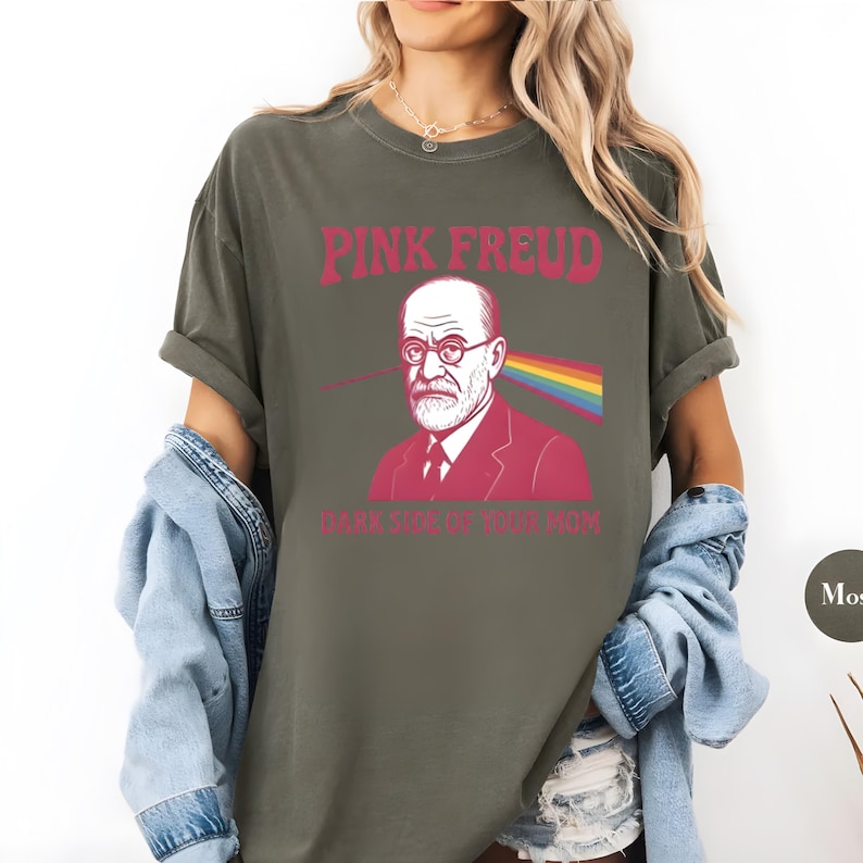 May include: Olive green t-shirt with the text "PINK FREUD" above an illustration of Sigmund Freud with a rainbow and the text "DARK SIDE OF YOUR MOM". The shirt has short sleeves and a relaxed fit.