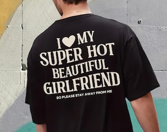 May include: Black t-shirt with white text on the back. The text reads "I ❤️ MY SUPER HOT BEAUTIFUL GIRLFRIEND SO PLEASE STAY AWAY FROM ME". The shirt is a casual style.