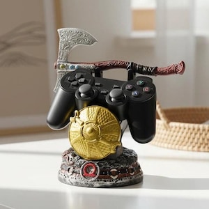 May include: A black games controller sits on a decorative stand. The stand features a gold shield, a silver axe, and a detailed base with red and grey accents. The controller is designed to hold the game controller.