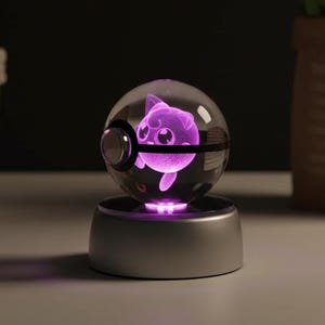 May include: A clear glass sphere with a purple-lit cartoon character inside, resting on a silver base. The sphere has a black band around it, resembling a Poké Ball. The background is dark, with a basket and a bouquet of pink tulips.