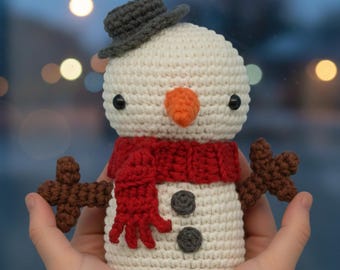 Peut inclure: Un bonhomme de neige crocheté avec un corps blanc, une écharpe rouge et un chapeau gris. Il a un nez en carotte, des yeux noirs et des bras en forme de bâton marron. Deux boutons gris sont sur le corps du bonhomme de neige.