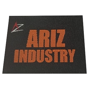 May include: A rectangular black fabric label featuring the embroidered text ARIZ INDUSTRY in bold orange capital letters. To the left of the text is a small stylized logo consisting of a red triangle shape above a white zigzag line.