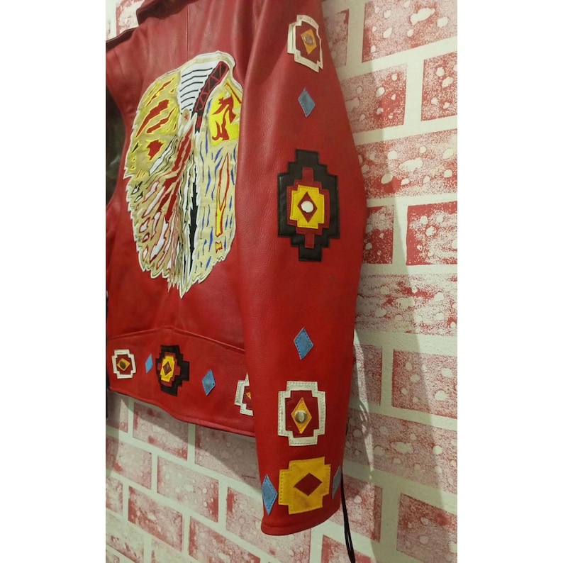 May include: A red leather jacket featuring a large, detailed graphic on the back depicting a stylized eagle in shades of yellow, white, and black. The sleeves and lower back are decorated with geometric patches in black, yellow, and white.