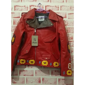 May include: A bright red motorcycle jacket featuring a dark brown inner collar, a silver zipper, and a hanging tag with the Indian Motorcycle logo. The jacket is decorated with yellow, brown, and blue geometric patches along the hem and sleeves.