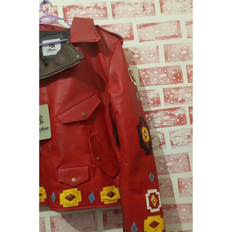 May include: A bright red leather jacket featuring a pointed collar, a dark grey inner lining, and two chest pockets with snap closures. The jacket is decorated with geometric patches in yellow, black, and blue, and includes a tag labeled Indian.