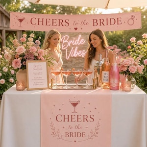 Könnte beinhalten: Ein rosafarbenes Tischläufer- und Banner-Set mit dem Text CHEERS to the BRIDE, Martini-Glas- und Ring-Symbolen sowie floralen Akzenten. Das Set enthält ein passendes Schild für Signature-Cocktails, gestaltet für eine feierliche Veranstaltung.