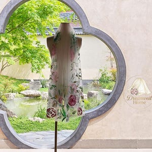 Puede incluir: Un velo blanco transparente con un estampado floral de rosas rosas y burdeos y hojas verdes. El velo se exhibe en un maniquí frente a una escena de jardín. El logotipo de Dreamveil Home es visible en la esquina inferior derecha.