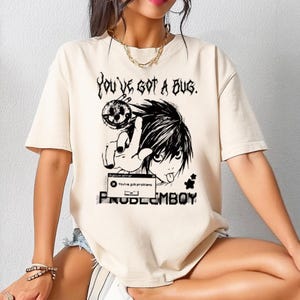 Puede incluir: Camiseta color crema con un gráfico en blanco y negro estilo anime. El gráfico incluye el texto "YOU'VE GOT A BUG" y "PROBLEMBOY", junto con una ilustración de personaje y un mensaje de error informático.