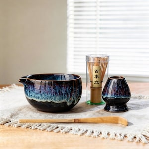 May include: A matcha tea set comprising a glazed ceramic bowl with a spout, a whisk, a whisk holder, and a bamboo scoop. The bowl and holder have a dark blue and purple glaze. The whisk is in a clear container.
