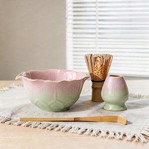 Peut inclure: Un bol à matcha en céramique avec un bord festonné, présentant un dégradé de rose tendre et de vert clair. À côté se trouvent un fouet en bambou, un porte-fouet en céramique assorti et une longue cuillère à thé en bambou incurvée.