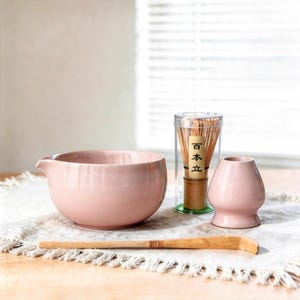 May include: A pink ceramic matcha bowl with a spout, a bamboo whisk in a clear container, a pink whisk holder, and a bamboo scoop. The items are arranged on a white cloth.