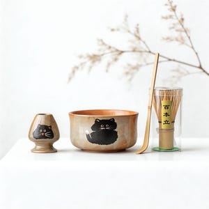 Könnte beinhalten: Ein Matcha-Teeset mit einer Keramikschale und einem Quirlhalter, beide mit schwarzen Katzenmotiven verziert. Ein Bambusbesen und -löffel sind ebenfalls enthalten. Für die Zubereitung von Matcha-Tee.