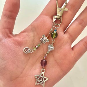 May include: A silver keychain with a lobster clasp, featuring a butterfly charm, a heart charm, and a star charm. The keychain has a spiral design and is adorned with colourful beads in shades of green, purple, and red.