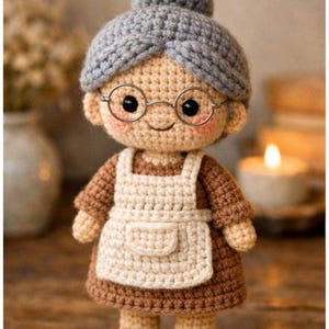 May include: A handmade crochet doll depicting a smiling elderly woman. The doll features a gray bun, round glasses, and a white apron over a brown dress. The doll is wearing beige shoes and has rosy cheeks.