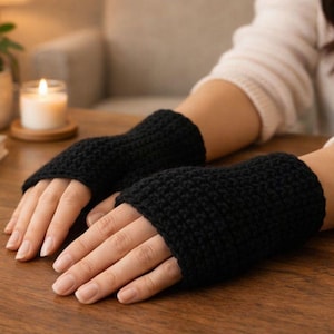 Puede incluir: Un par de guantes sin dedos negros, tejidos a ganchillo con un punto texturizado. Los guantes cubren las muñecas y se extienden hasta los nudillos, dejando los dedos libres. Los guantes se exhiben sobre una superficie de madera.