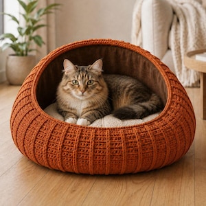 May include: A brown tabby cat rests inside a round, orange crochet pet bed. The bed has a soft, brown interior and a cream-colored cushion. The cat is looking at the camera. The bed is on a wooden floor.