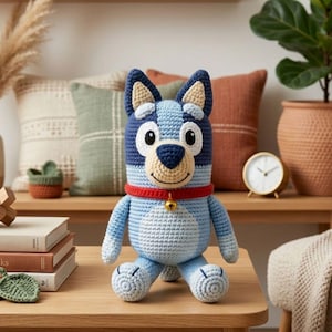 May include: A handmade crochet dog doll in shades of blue, navy, and cream, with a red collar and a small gold bell. The plush toy has large white eyes and a smiling face. The dog is sitting on a wooden surface.