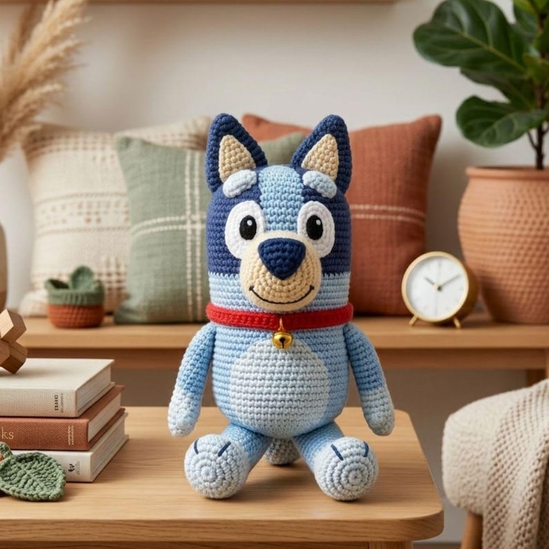 May include: A handmade crochet dog doll in shades of blue, navy, and cream, with a red collar and a small gold bell. The plush toy has large white eyes and a smiling face. The dog is sitting on a wooden surface.