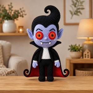 May include: A crocheted vampire doll with light purple skin, large orange eyes, and two white fangs. The doll has black hair styled in a tall curl, a black cape with a red interior lining, a white shirt, and black pants, all crafted with yarn.