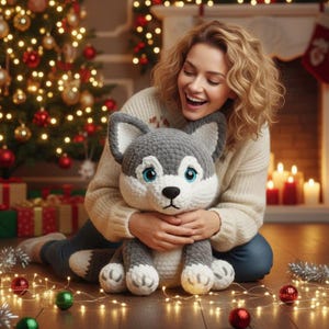 May include: A handmade gray and white husky dog plush toy with blue eyes. The plush toy is being held by a person in a cream-colored sweater. The background includes a Christmas tree, fireplace, and holiday decorations.