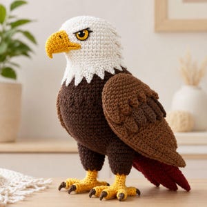 Puede incluir: Una figura de águila calva tejida a ganchillo con cabeza blanca, pico amarillo brillante y plumas corporales de color marrón oscuro. Las alas tienen textura de hilo marrón en capas y el ave se sostiene sobre dos patas amarillas con garras.