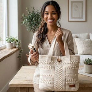 Könnte beinhalten: Cremefarbene Häkeltasche mit Holzknopfverschluss. Die Tasche hat ein strukturiertes Muster mit Noppen und Zickzack-Designs. Die Tasche hat stabile Griffe und ein kleines braunes Lederetikett. Die Tasche wird von einer Person gehalten.