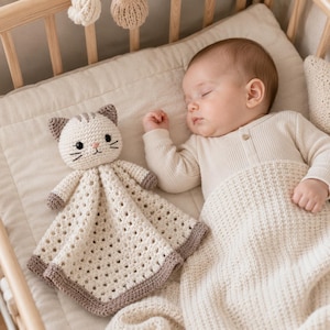 May include: A cream-colored crochet cat security blanket featuring a small stuffed cat head with brown ears, embroidered black eyes, and whiskers. The blanket body has a square, open-stitch pattern with a solid brown border along the bottom edge.