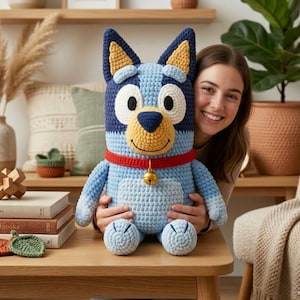 May include: A large, handmade crochet dog plushie in shades of blue, yellow, and white. The dog has a red collar with a small gold bell. The plushie is sitting on a wooden table.